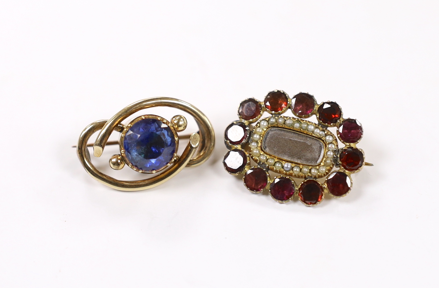 A Victorian yellow metal and single stone foil backed sapphire set scrolling mourning brooch, verso with hair beneath a glazed panel, 26mm, together with a Regency yellow metal, garnet and seed pearl cluster set mourning